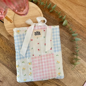 May include: A patchwork book sleeve with a gingham pattern in blue and pink. The sleeve has a white ribbon tie and a label that reads "BOOK CLUB". The white fabric is decorated with floral accents.