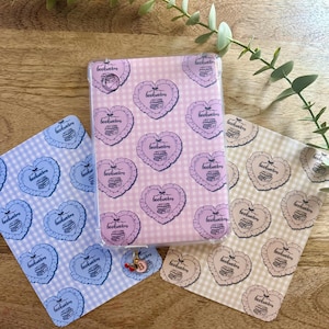 May include: A clear phone case featuring a pink gingham pattern with heart-shaped "bookworm" designs. Two matching cards in blue and beige are also visible, along with a charm.
