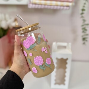 May include: A clear glass tumbler with a bamboo lid and a stainless steel straw. The tumbler is decorated with pink hydrangea flowers and green leaves. The glass is filled with a light brown liquid.