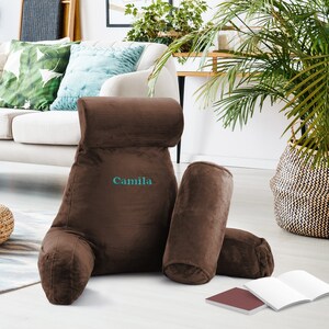 May include: A brown plush reading pillow with a back support and two armrests. The pillow has the name "Camila" embroidered on the back.
