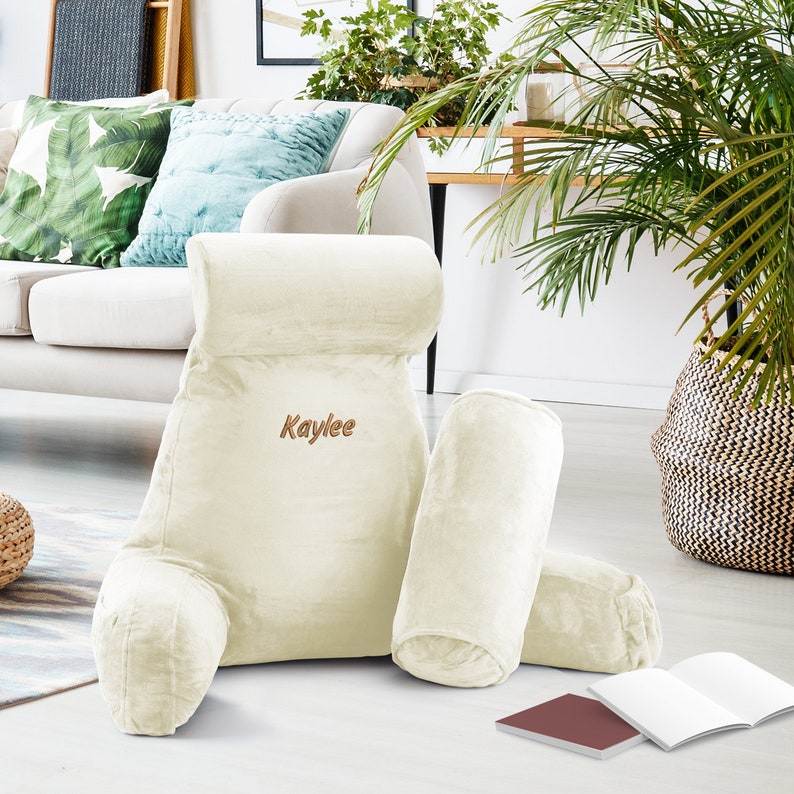 May include: A large, cream-coloured reading pillow with a smaller pillow and a lumbar pillow. The reading pillow has the name "Kaylee" embroidered on it.