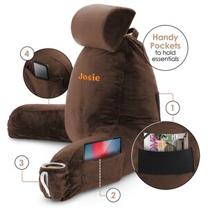May include: A brown plush reading pillow with a headrest and multiple pockets. The pillow has a personalised name "Josie" embroidered on the front. The pockets are labelled with numbers 1 through 4 and are designed to hold items like a phone, tablet, or a book.