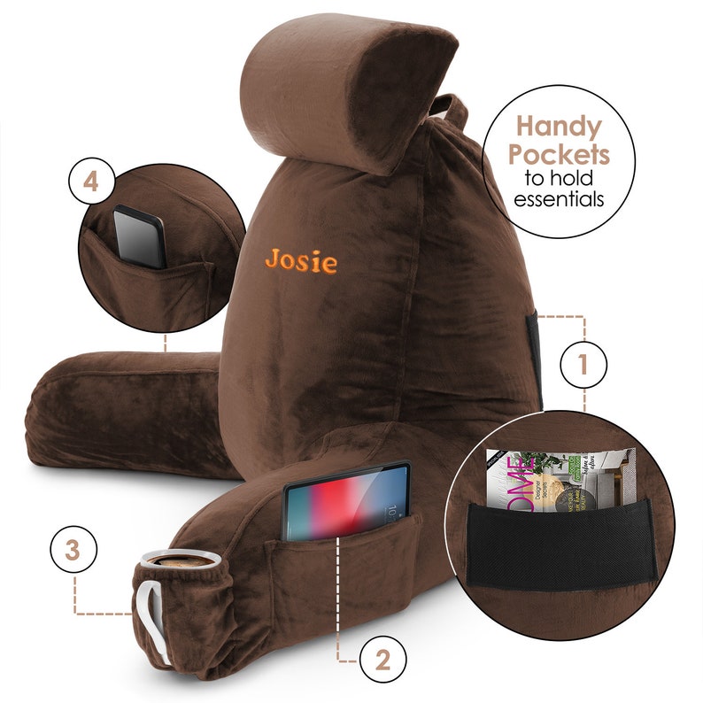 May include: A brown plush reading pillow with a headrest and multiple pockets. The pillow has a personalised name "Josie" embroidered on the front. The pockets are labelled with numbers 1 through 4 and are designed to hold items like a phone, tablet, or a book.