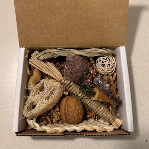 May include: A cardboard box filled with a variety of small pet enrichment items, including a whole walnut, a loofah sponge, a braided straw mat, a woven rattan ball, a dried flower sphere, a peanut, and several bundles of natural grass and wood sticks.