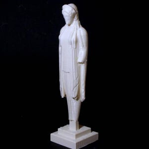 Könnte beinhalten: Eine weiße Statue einer bekleideten Figur, die auf einem quadratischen Sockel steht. Die Figur hat ein drapiertes Gewand und eine Kopfbedeckung. Die Statue steht vor einem schwarzen Hintergrund.