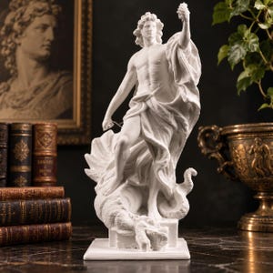 Könnte beinhalten: Eine weiße Skulptur einer Figur, die auf einer gewundenen Schlange steht und einen Gegenstand in einer Hand in die Höhe hält. Die Figur ist in fließenden Stoff gehüllt, der sich um die Taille und über die Schulter legt, auf einem quadratischen Sockel.