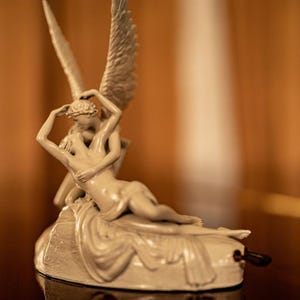 Puede incluir: Una escultura color crema que representa dos figuras abrazadas, una con grandes alas. Las figuras están entrelazadas, una reclinada y la otra inclinada. La escultura está sobre una base con una pequeña manivela.