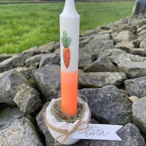 Könnte beinhalten: Eine weiße Kerze mit Karottenillustration und orangefarbenem Farbverlauf. Die Kerze steht in einem weißen, eiförmigen Halter mit Stroh und Bindfaden. Ein Anhänger mit "Happy Easter" ist angebracht.