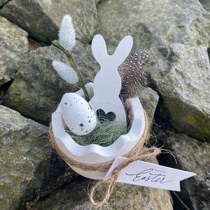 Könnte beinhalten: Weiße, eierschalenförmige Osterdekoration mit einem Holzhase-Ausschnitt, einem gesprenkelten Ei und Federn. Die Dekoration ist mit grünem Moos gefüllt und mit Bindfaden umwickelt. Ein kleines Etikett trägt die Aufschrift "Frohe Ostern."