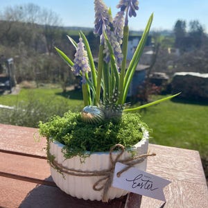Könnte beinhalten: Ein weißer, gerippter Keramiktopf mit grünem Moos, lila und weißen Blumen und einem dekorativen Ei. Ein kleines Etikett, das mit Bindfaden befestigt ist, lautet "Frohe Ostern". Der Topf steht draußen auf einem Holztisch.