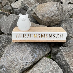 Könnte beinhalten: Weiße Keramik-Bärenfigur mit einem Herz auf der Brust, die auf einem Holzregal mit dem eingravierten Wort "HERZENSMENSH" steht. Das Regal liegt auf einem Stapel grauer Steine.