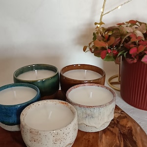 Può includere: Cinque candele in ceramica di vari colori: blu, verde, marrone e bianco maculato. Ogni candela ha un interno in cera bianca e un'unica stoppino. Le candele sono disposte su una superficie di legno, con un vaso rosso e del fogliame sullo sfondo.