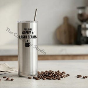 Puede incluir: Un vaso de acero inoxidable plateado con tapa transparente y pajita metálica. El vaso tiene el texto "FUELED BY COFFEE & LASER BEAMS" con una taza de café y un rayo. Granos de café esparcidos alrededor.
