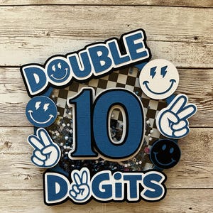 May include: A blue and white cake topper with the words "DOUBLE 10 DIGITS". The number 10 is in the center, surrounded by smiley faces and peace signs. The background has a checkered pattern.