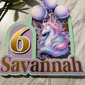 May include: Purple and green cake topper with a unicorn, balloons, and the number 6. The name Savannah is written in gold letters.
