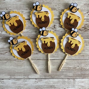 May include: Six yellow and white honey pot cupcake toppers with a bee on top. Each topper has the word "Hunny" written on it.