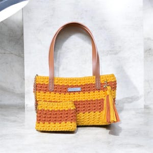 May include: A crocheted handbag and matching small pouch featuring horizontal stripes of mustard yellow and burnt orange yarn. The bag has two tan leather handles, a silver metal brand tag, and a decorative tassel made of matching yarn.