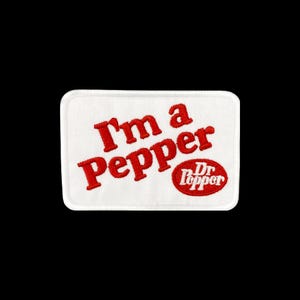 May include: A rectangular white embroidered patch featuring the red text I'm a Pepper in a slanted font, accompanied by a small red oval containing the white Dr Pepper logo. The patch has a clean white border and a textured fabric surface.