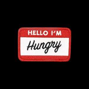 May include: A rectangular embroidered patch with a red border and a white centre. The top red section features the white text HELLO I'M in a bold sans-serif font. The white centre section features the black text Hungry in a cursive script font.