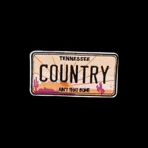 Op de afbeelding: Een rechthoekige geborduurde patch met een perzikkleurige achtergrond en een zonnestraalontwerp. De tekst TENNESSEE COUNTRY AIN'T THAT SOME staat in het zwart. Een roze cactus en een klein roze silhouet van een bokkend paard zijn onderaan zichtbaar.