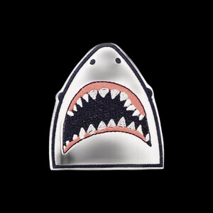Op de afbeelding: Een geborduurde patch in de vorm van een haaienkop. De patch heeft een wit bovengedeelte met twee kleine zwarte stippen als ogen, een wijd open mond met roze tandvlees en scherpe witte tanden tegen een donkerblauwe achtergrond, omlijnd in marine.