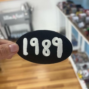 Peut inclure: Écusson ovale noir avec l'année "1989" brodée en blanc. L'écusson est présenté sur un fond d'intérieur de magasin avec des étagères et des articles.