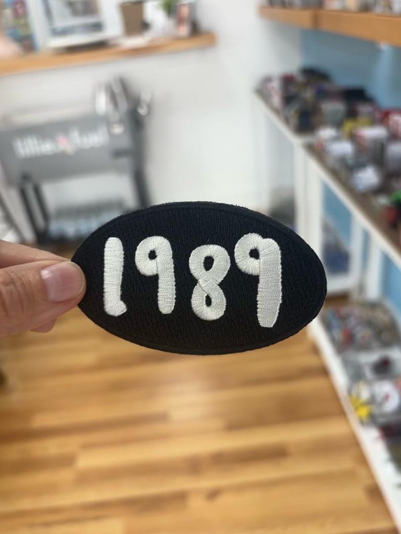 May include: Black oval patch featuring the year "1989" in white embroidery. The patch is held up against a shop interior with shelves and various items.