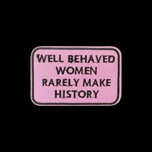 May include: A rectangular embroidered patch with a light pink background and a black border. The text WELL BEHAVED WOMEN RARELY MAKE HISTORY is stitched in black capital letters across the centre of the patch in a simple, bold font.