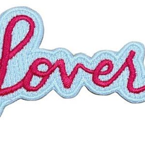 Op de afbeelding: Geborduurde patch met het woord "Lover" in een sierlijk lettertype. De roze tekst staat op een lichtblauwe achtergrond, met een lichtblauwe omtrek en een geschulpte rand. Geschikt voor kleding.