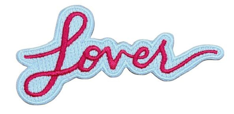 Op de afbeelding: Geborduurde patch met het woord "Lover" in een sierlijk lettertype. De roze tekst staat op een lichtblauwe achtergrond, met een lichtblauwe omtrek en een geschulpte rand. Geschikt voor kleding.