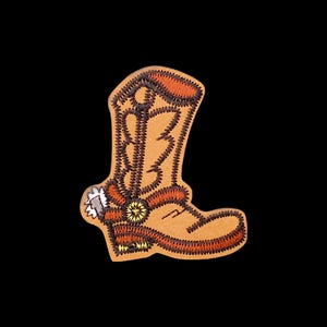 May include: A tan embroidered patch in the shape of a cowboy boot. The design features dark brown stitching for the boot outline and decorative patterns, with a silver-colored spur attached to the heel area and orange accents on the top.