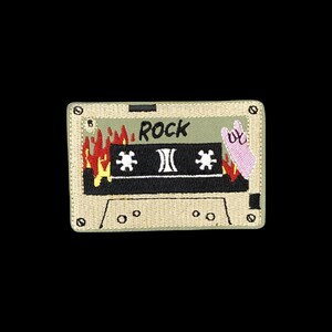 May include: A rectangular embroidered patch shaped like a cassette tape. The design features a light green background, a black centre strip with white spools, red and yellow flames, the word ROCK in black text, and a pink hand making a rock sign.