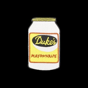 May include: An embroidered patch shaped like a jar of Duke's Mayonnaise. The patch features a white jar with a yellow label, a black oval containing the yellow text Duke's, and the word MAYONNAISE in orange letters below the oval.