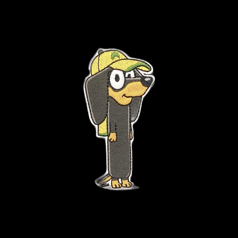 May include: An embroidered patch featuring a cartoon dog with long dark ears, a tan muzzle, and a yellow baseball cap with a green brim. The dog has large white eyes, a black nose, and is depicted in a standing position with tan paws.