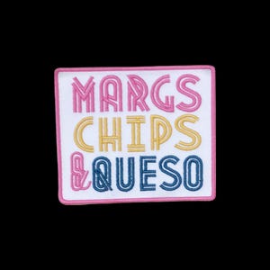 May include: A rectangular embroidered patch with a white background and a pink border. The text reads MARGS in pink, CHIPS in yellow, and & QUESO in blue. The lettering features a double-line style, and the patch has a clean, stitched edge.