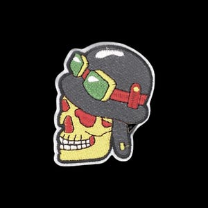May include: An embroidered patch featuring a yellow skull wearing a dark grey helmet. The helmet is adorned with green-tinted goggles that have a red strap. The design is outlined in white thread, creating a distinct shape against the black background.