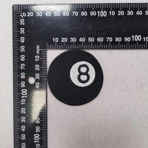 May include: A circular black embroidered patch featuring a white center circle with the black number 8, positioned next to a black metal L-shaped ruler showing metric measurements, with the patch diameter measuring approximately 50 millimeters.