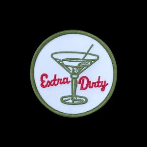 Puede incluir: Un parche bordado circular con fondo blanco y borde verde oliva. El diseño presenta una copa de martini verde oliva con un agitador y el texto bordado en rojo Extra Dirty escrito a través del centro de la copa de martini.
