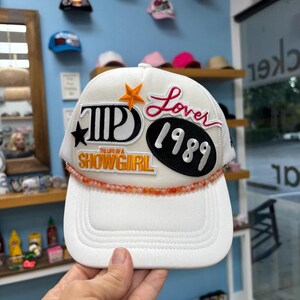 May include: White trucker hat with a mesh back and a curved brim. The hat features embroidered patches with the words "Lover 1989" and "The Life of a Showgirl". A beaded orange strand accents the brim.