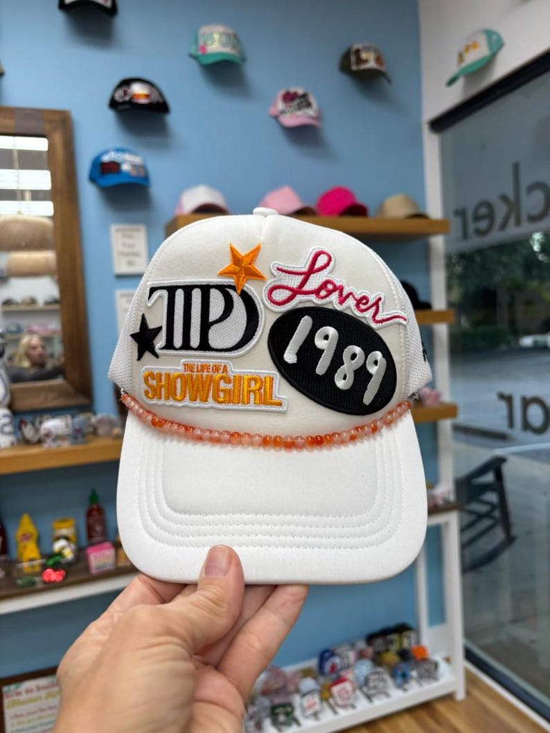 May include: White trucker hat with a mesh back and a curved brim. The hat features embroidered patches with the words "Lover 1989" and "The Life of a Showgirl". A beaded orange strand accents the brim.