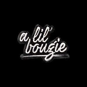 Op de afbeelding: Een geborduurde patch met de witte cursieve tekst a lil bougie op een zwarte achtergrond. De belettering is gestileerd met een dikke witte rand en een horizontale lijn onder het woord bougie, wat een opvallend grafisch ontwerp creëert.