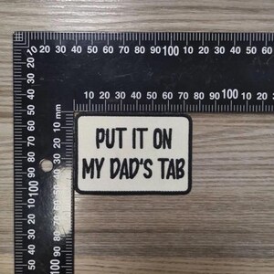 May include: A rectangular embroidered patch with a white background and a black border. The text "PUT IT ON MY DAD'S TAB" is stitched in black capital letters. A black metal ruler is positioned along the top and left edges for scale.