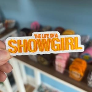 May include: Embroidered patch with the text "THE LIFE OF A SHOWGIRL" in orange letters on a white background. The patch is rectangular with a curved top and bottom. The patch is held in a person's hand.