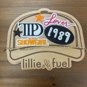 May include: A wooden display featuring the text "Lover 1989" and "The Life of a Showgirl". A gold-coloured necklace is draped across the display. The display has an orange star and a black star.