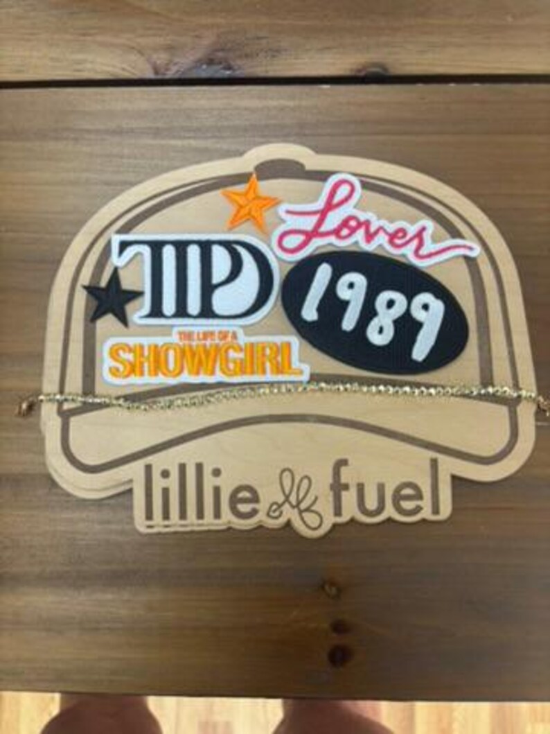May include: A wooden display featuring the text "Lover 1989" and "The Life of a Showgirl". A gold-coloured necklace is draped across the display. The display has an orange star and a black star.