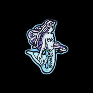 Può includere: Una toppa ricamata raffigurante una sirena con lunghi capelli fluenti viola e bianchi. La figura ha una coda azzurra con un motivo a scaglie e un top a forma di conchiglia. L'intero disegno è delineato da un bordo spesso di colore azzurro.