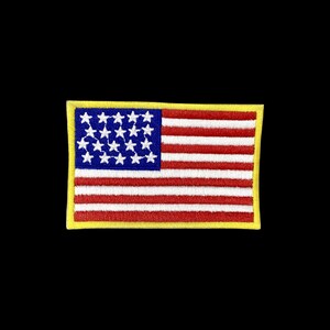 Puede incluir: Un parche bordado rectangular con el diseño de la bandera de los Estados Unidos y un borde amarillo. El cantón azul contiene veinticuatro estrellas blancas dispuestas en filas, y el campo consta de franjas horizontales rojas y blancas alternas.