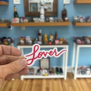 Op de afbeelding: Een witte patch met het woord "Lover" geborduurd in rood cursief schrift. De patch wordt vastgehouden voor een winkeldisplay met diverse items op planken en tafels.