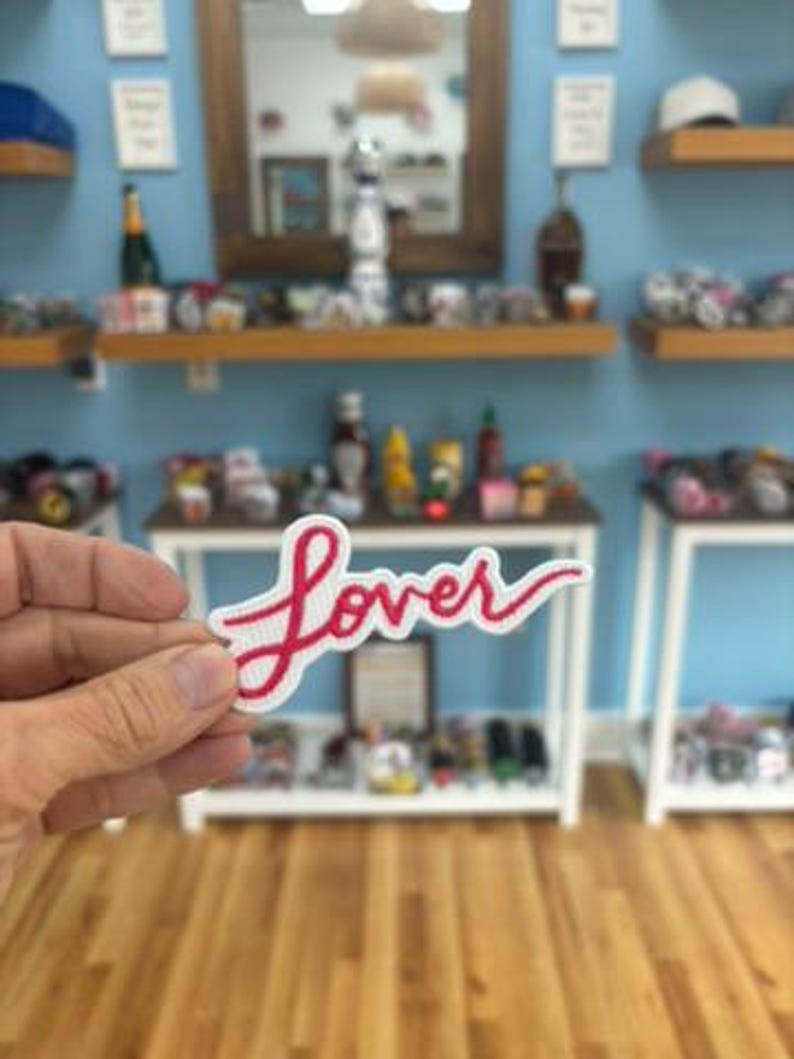 Op de afbeelding: Een witte patch met het woord "Lover" geborduurd in rood cursief schrift. De patch wordt vastgehouden voor een winkeldisplay met diverse items op planken en tafels.