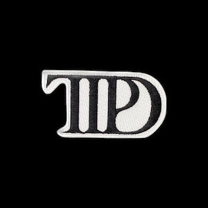 May include: An embroidered patch featuring a stylised black and white design. The graphic consists of a bold black letter D integrated with vertical black stripes on a white background, all enclosed within a rounded white border outline.